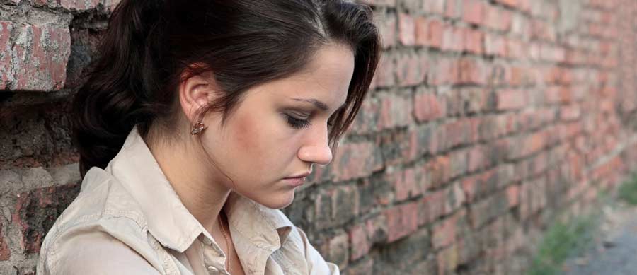 Serious young woman leaning against a brick wall, head bowed and reflecting, illustrating the emotional weight of recognizing one’s need for drug or alcohol rehab.