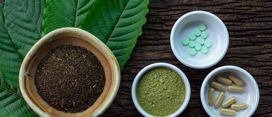 kratom addiction- Various forms of kratom including dried leaves, powdered kratom, capsules, and tablets displayed on natural wood background with green kratom leaves.