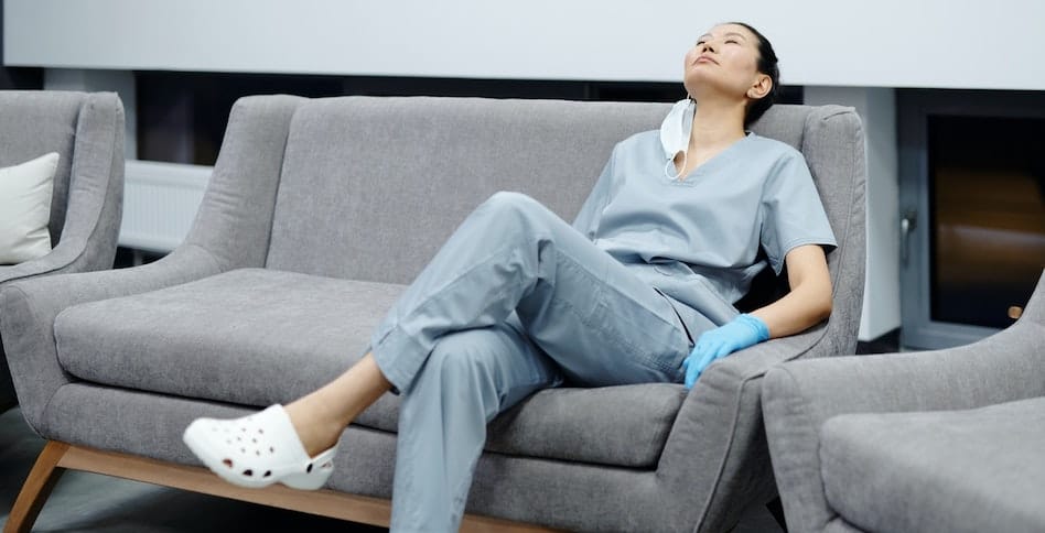 Exhausted medical professional in scrubs resting on a couch, highlighting the stress that can lead to addiction in healthcare workers.