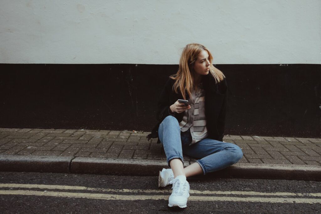 Young woman sitting on the curb with a phone, reflecting on the need for professional help in drug addiction rehab.
