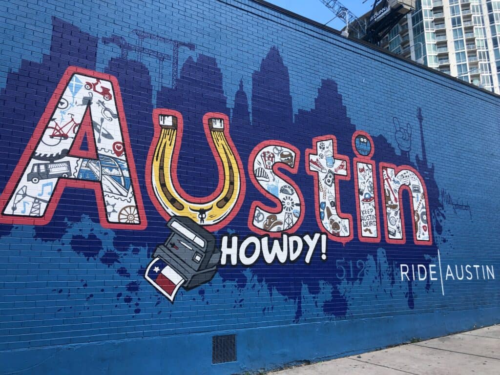 Colorful mural on a brick wall spelling ‘Austin Howdy!’ with illustrated icons like a horseshoe, skyline, bike, and ‘Keep Austin Weird’ motifs.