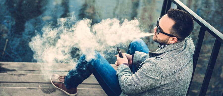 Man vaping outdoors releasing large clouds of vapor, illustrating the addictive nature of e-cigarettes and nicotine use.